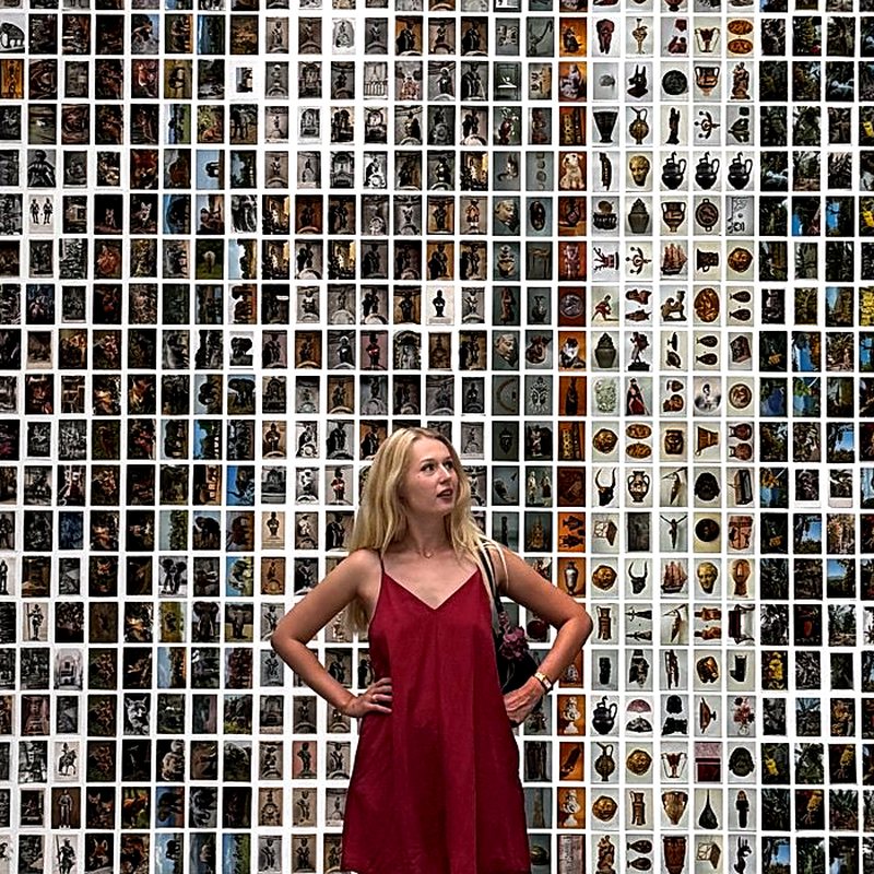 Julia standing in a room whose walls are covered with thousands of small framed pictures